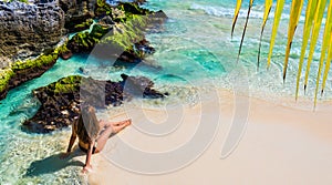 Young fashion woman in bikini sitting on tropical beach. Beautiful girl in black swimsuit stands on the shore. Caribbean