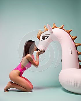 Young fashion woman kissing big inflatable unicorn pegasus float mattress in pink bikini for swimming pool