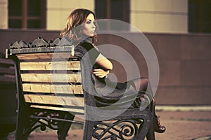 Young fashion woman in long sleeveless coat sitting on bench