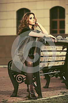 Young fashion woman in long sleeveless coat sitting on bench