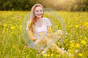 Young fashion woman posing in summer meadow. Trendy girl at nature landscape background. Happy smiling female sitting among yellow