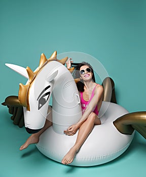 Young fashion woman relaxing with big inflatable unicorn pegasus float mattress in pink bikini for swimming pool