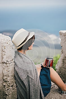 Young fashion woman sitting on a top of the mountain drinking tea with beautiful mountains and ocean view on the background