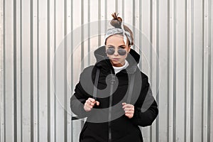 Young fashionable beautiful hipster woman with a stylish hairstyle with a bandana in black sunglasses in a long black coat with a