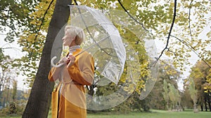 Young fashionable blond caucasian woman with transparent umbrela looking around in the park in autumn