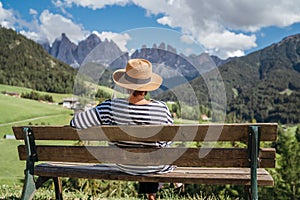 Young fashionable dressed female in straw hat sitting on a bench enjoying Santa Maddalena village view and stunning picturesque