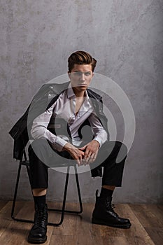 Young fashionable hipster man in a stylish black leather jacket in a white classic shirt in jeans in trendy boots sits on a