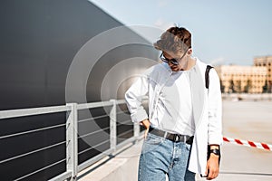 Young fashionable man model in a T-shirt in a white shirt in vintage blue jeans in sunglasses walks around the city
