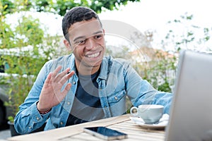 Young fashionable man sitting in cafe with laptop waving