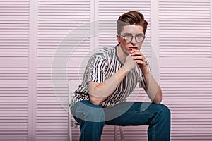 Young fashionable man with a stylish hairstyle in a fashion striped T-shirt in vintage glasses posing on a chair near a wooden