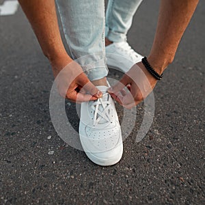 Young fashionable manstraightens laces on trendy white leather sneakers. New stylish collection of men`s shoes. Summer fashion.