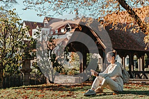 Young Fashionable Teenage Girl With Smartphone In Europian Park In Autumn Sitting At Smiling. Trendy Young Woman In Fall