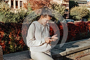 Young Fashionable Teenage Girl With Smartphone In Europian Park In Autumn Sitting At Smiling. Trendy Young Woman In Fall