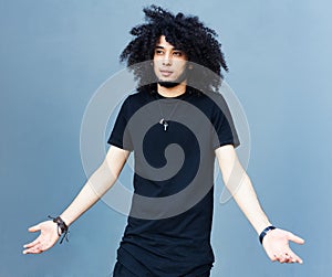 A young fashionably dressed man with an afro hairstyle and a beard in a cool outfit posing on a gray wall. He spreads his arms sho