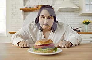 Young fat excited woman sitting at the table in kitchen looking at the plate with big burger.