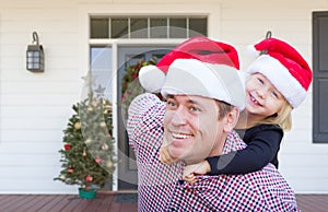 Young Father and Daughter Wearing Santa Hats On Front Porch of House With Christmas Decorations