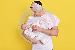 Young father feeding his baby while posing isolated over yellow background, wearing white t shirt and sleeping mask on forehead,