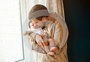 Young father gently holding and kissing his baby near the window at home