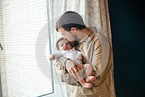 Young father gently holding and kissing his baby near the window at home