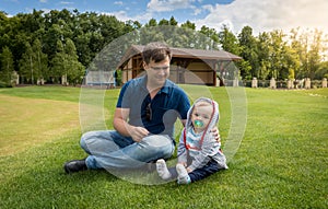 Young father with his baby son relaxing on grass at park at sunny day