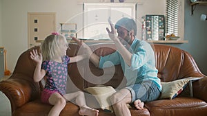 A young father and his cute happy little daughter are giving each other high fives sitting on the sofa. Slow motion