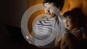 Young father and his little son watching cartoon movie using tablet computer and talking while lying in bed in evening