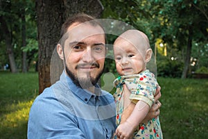 Young father holding a baby son on the background of a summer landscape. Bearded man smiling and happy, looking at camera