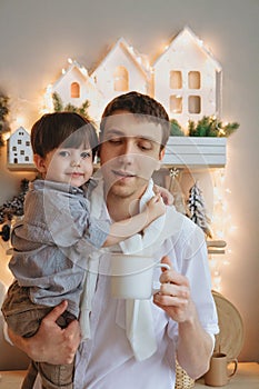 Young father holding cute little boy and cup of cacao, spending time with son on Christmas at home