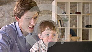 Young father holding his smiling son and looking at laptop, sitting in modern office, happy