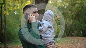 A young father holds his son in his arms and talks on the phone. A man with a child in the park with a smartphone.