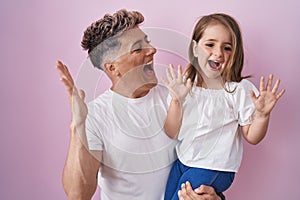 Young father hugging daughter over pink background celebrating victory with happy smile and winner expression with raised hands