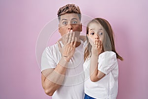 Young father hugging daughter over pink background covering mouth with hand, shocked and afraid for mistake