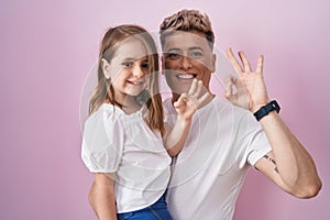 Young father hugging daughter over pink background doing ok sign with fingers, smiling friendly gesturing excellent symbol