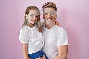 Young father hugging daughter over pink background looking positive and happy standing and smiling with a confident smile showing