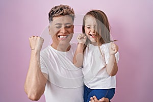 Young father hugging daughter over pink background screaming proud, celebrating victory and success very excited with raised arms