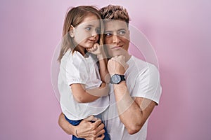 Young father hugging daughter over pink background serious face thinking about question with hand on chin, thoughtful about
