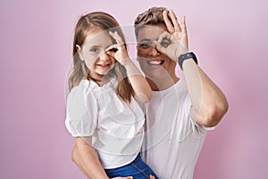 Young father hugging daughter over pink background smiling happy doing ok sign with hand on eye looking through fingers