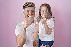 Young father hugging daughter over pink background smiling happy and positive, thumb up doing excellent and approval sign