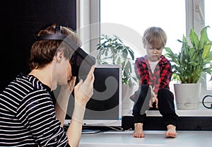 A young father plays in a virtual reality helmet. Little boy son is sitting nearby and crying. Child needs attention