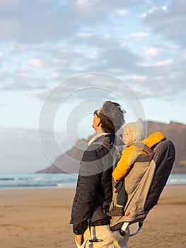 Young father rising hands to the sky while enjoying pure nature carrying his infant baby boy son in backpack on windy