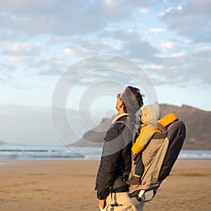 Young father rising hands to the sky while enjoying pure nature carrying his infant baby boy son in backpack on windy