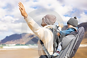 Young father rising hands to the sky while enjoying pure nature carrying his infant baby boy sun in backpack on windy