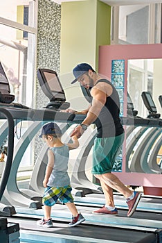 Young father running on racetrack in spacy gym with little son.