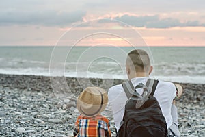 Young father and son are sitting on the beach against the backdrop of the sea and the setting sun