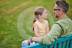 Young father spending time with his cute little daughter. They are looking at each other while sitting on the bench in