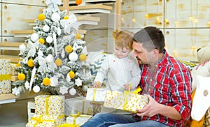 Young father and toddler daughter sitting near Christmas tree and opening gift boxes