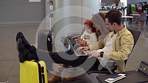 Young father working on laptop on airport with his family while waiting for departure. Long haired mother entertains her