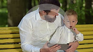 A young father and a young son are sitting on a park bench and playing on a tablet. Father`s Day.