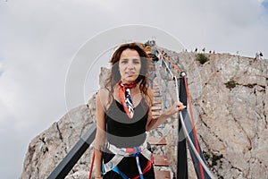 A young fearless woman in safety gear walks over a rope bridge over a precipice high in the mountains.