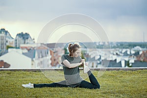 Young female acrobat on the grass performs twine, cityscape on background